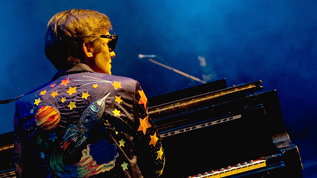 a man dressed as Elton John is seen from behind and sitting at a piano
