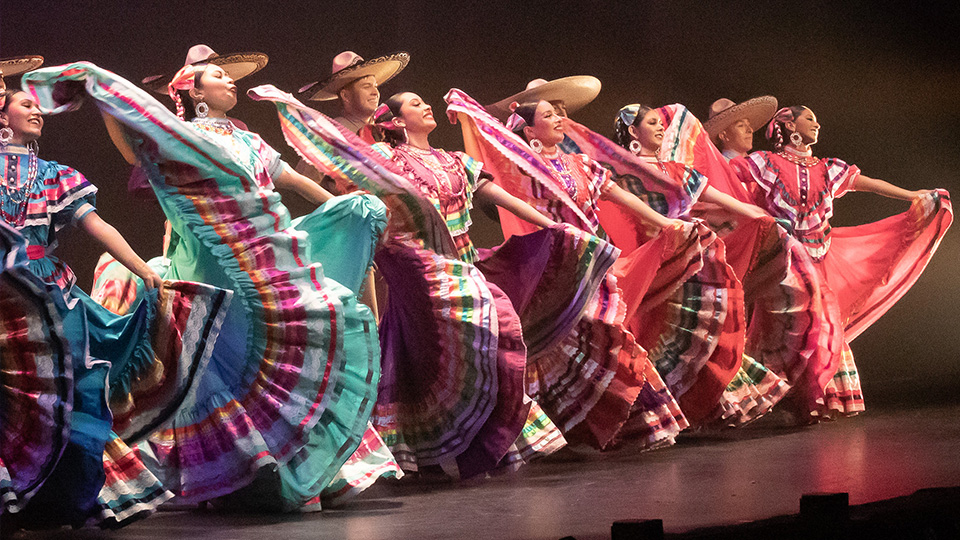 six dancers in colorful, traditional dresses lift their skirts in a semi-circle