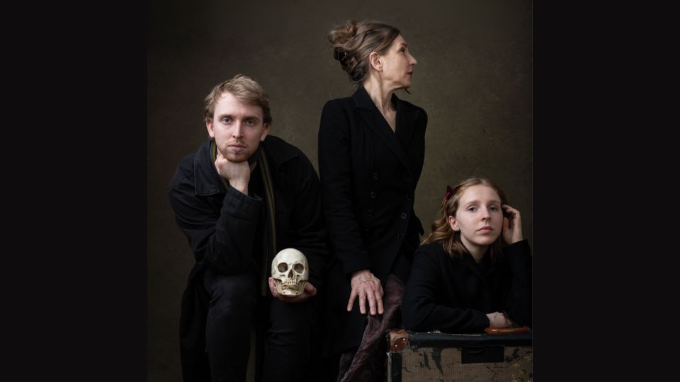 Three actors in black pose with a skull for a studio portrait against a dark backdrop.