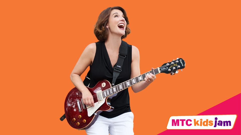 Krista Tatschl Eyler smiles while holding a guitar. She is standing against an orange background.