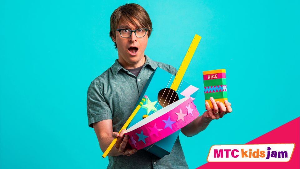 Justin Roberts holding brightly colored paper crafts in the shapes of instruments. He is standing against a bright blue background.