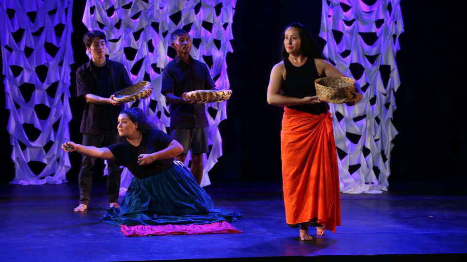 performers on stage - a woman carrying a basket walks past three other people, two of whom are standing holding baskets and another who is on her knees with arms outstretched