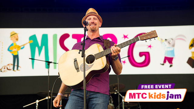 Ben Gundersheimer on stage with a guitar and a banner behind him that reads Mister G