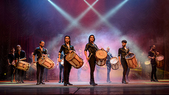a group of men stand in formation on a stage lit with spotlights and fog while holding large drums