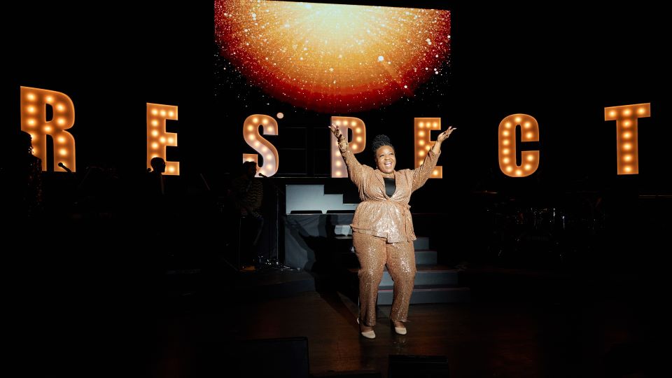 an Aretha Franklin impersonator stands on stage with her hands upraised in front of lit up letters that spell out respect