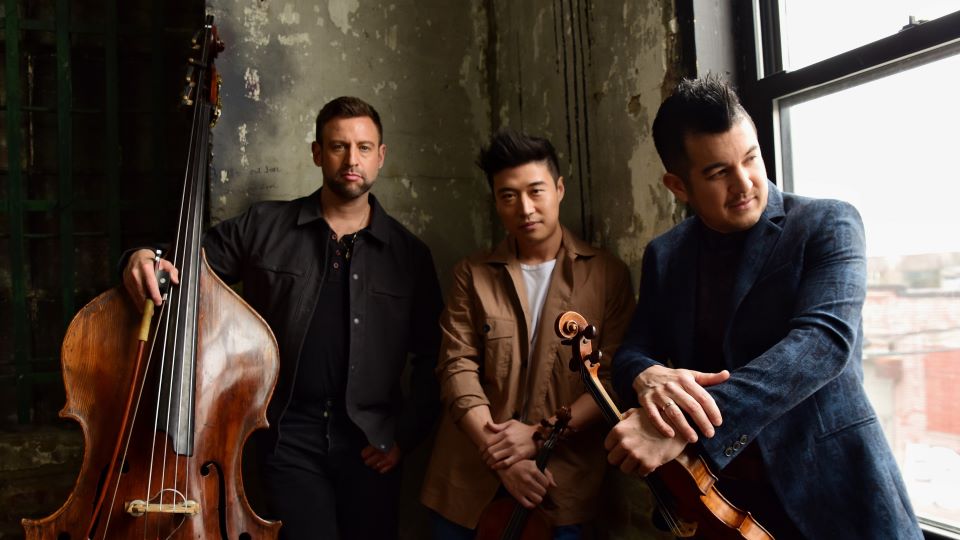 The three members of Time for Three pose for a picture next to a large picture window. They are wearing suits and holding their instruments. They are bathed in a soft light.
