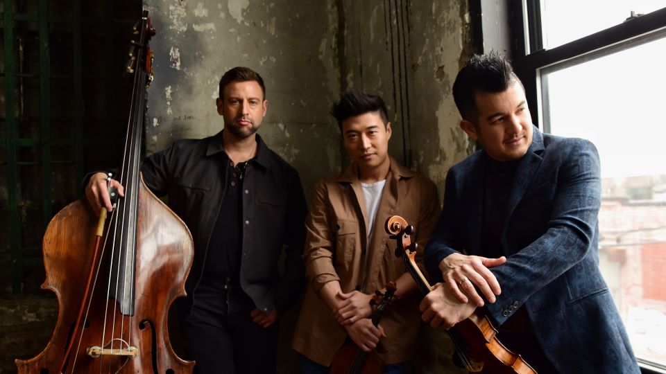 The three members of Time for Three pose for a picture next to a large picture window. They are wearing suits and holding their instruments. They are bathed in a soft light.