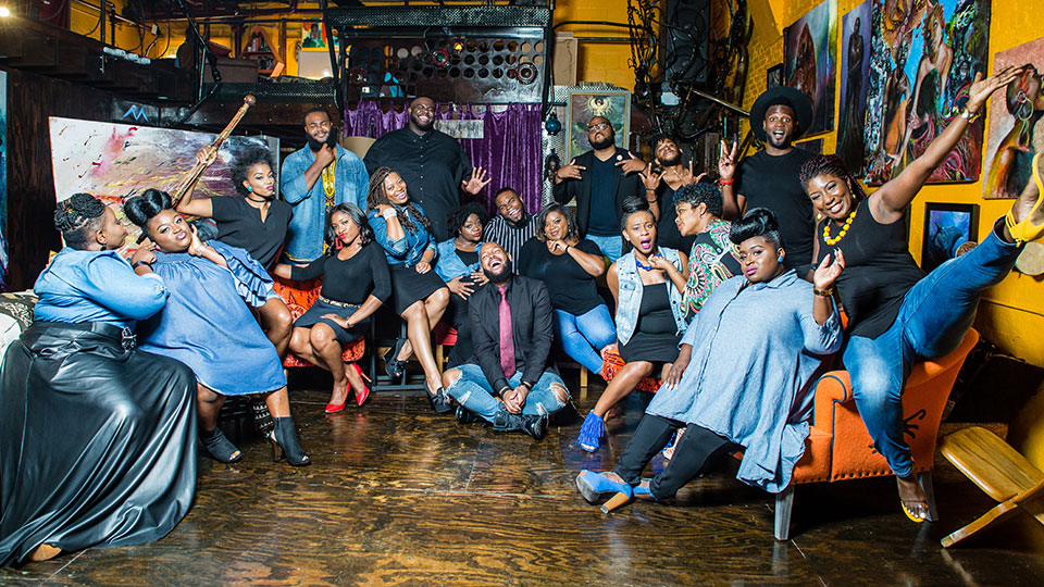 a large, celebratory group of musicians poses in an art studio with yellow walls and many art works surrounding them