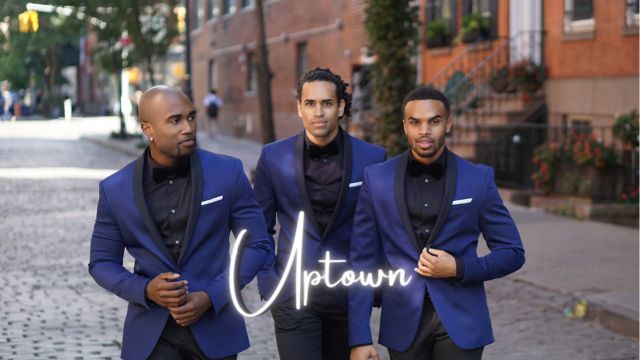 The three members of Uptown walk down a street next to a brick building. All three men are wearing blue jackets.