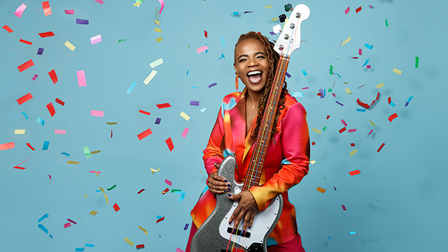 A smiling Divinity Roxx stands holding her guitar while confetti falls all around her.
