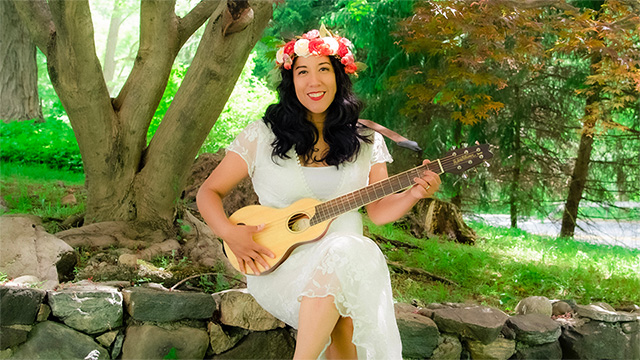 Flor Bromley sitting on a rock wall in a park. She is wearing a white dress, a flower hat and is playing a stringed instrument.