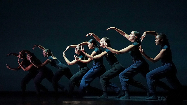 dancers in a line on a dark stage, each with their arms outreached in a circular shape