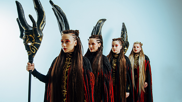 The four members of Yagody standing in a line. They are wearing cloaks, their hair is braided in rows, and they are holding ornate masks mounted to the top of long poles.