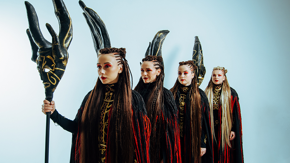 The four members of Yagody standing in a line. They are wearing cloaks, their hair is braided in rows, and they are holding ornate masks mounted to the top of long poles.