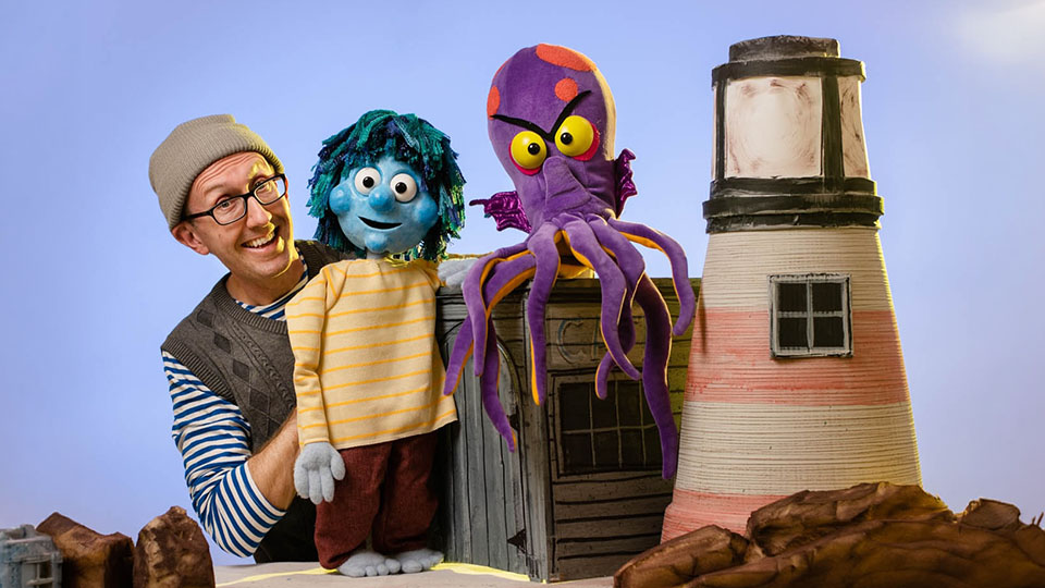 a smiling man next to two puppets and a model lighthouse