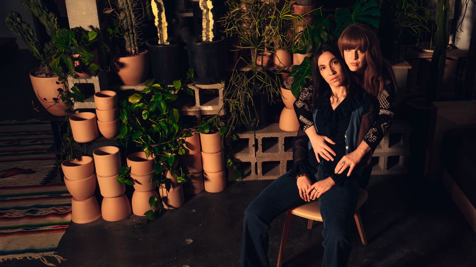 Two women next to several houseplants and terracotta pots. One woman is standing behind and has her arms around the other woman who I seated in a chair. They both look at the viewer with a serious expression.