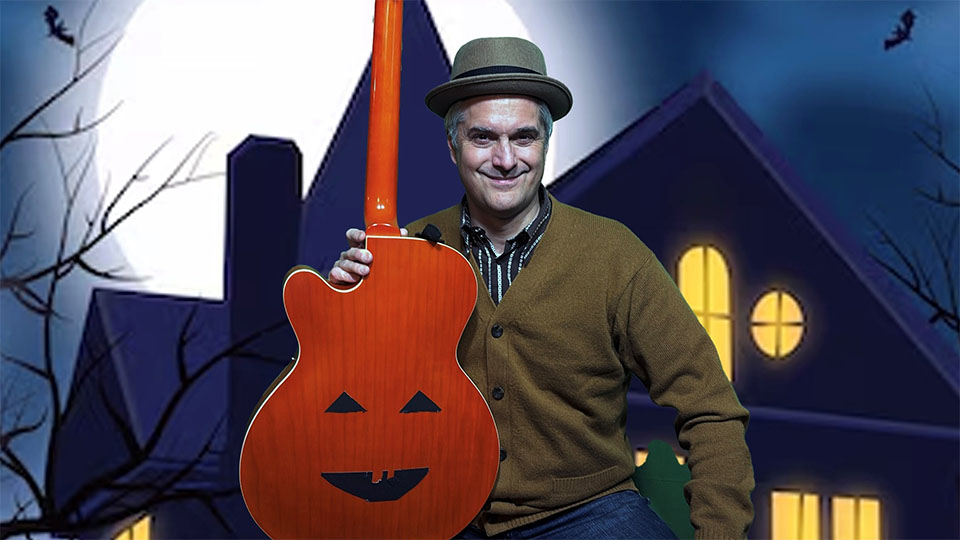 Matt Heaton stands in front of a haunted house and full moon while holding a pumpkin guitar