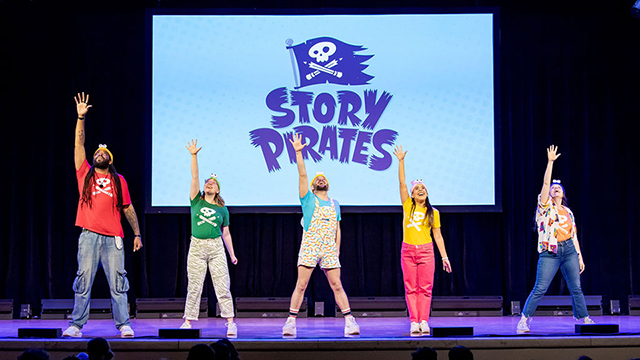 5 people standing on a stage with one arm raised are in front of a backdrop that says Story Pirates
