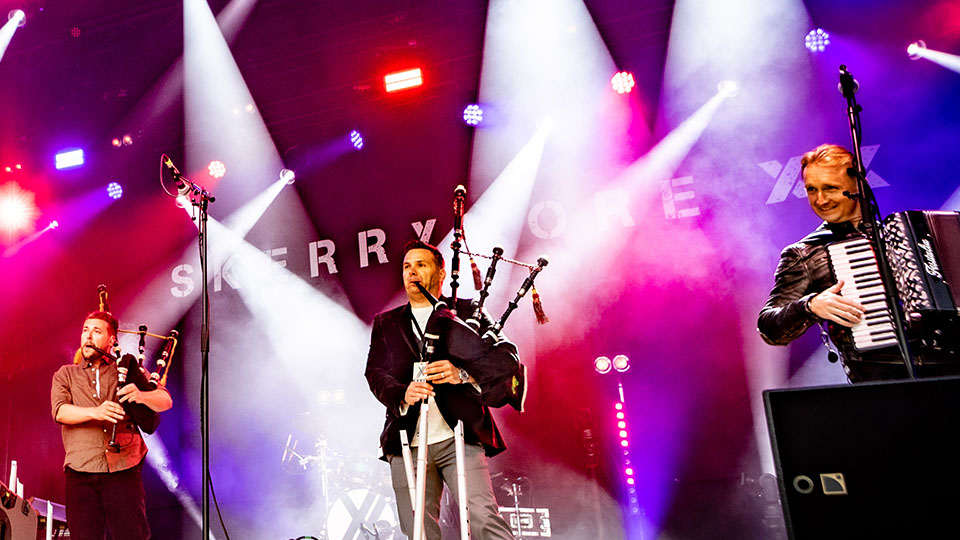 two bagpipers and an accordion player perform on a stage lit by bright colors and spot lights