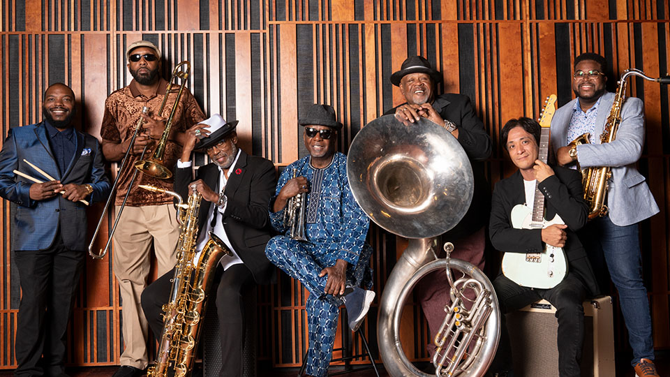 7 band members lined up against a studio wall. They are holding their respective instruments and smiling at the viewer.