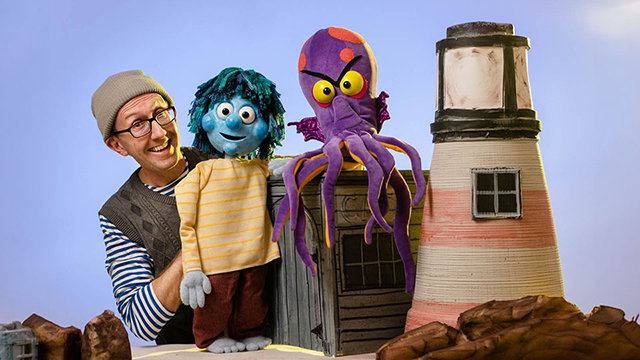 a smiling man next to two puppets and a model lighthouse