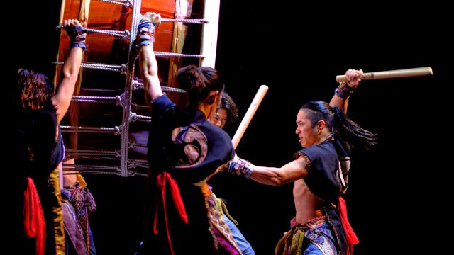 a man pounds a huge drum that is being held up by four other people