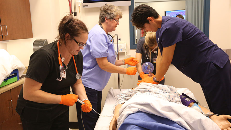 Three students practices aspects of healthcare on a simulation mannequin while an instructor looks on.