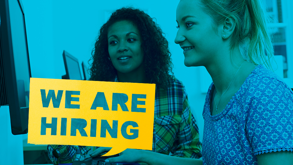 Two smiling women stand next to a We're Hiring sign.