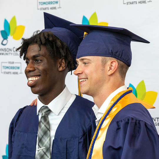 Two graduates pose for a photo in front of a backdrop that has JCCC logos on it