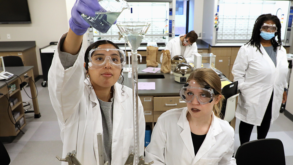 two jccc students in lab coats and safety gear pour a beaker of liquid into a tall funnel while other students work in the lab in the background