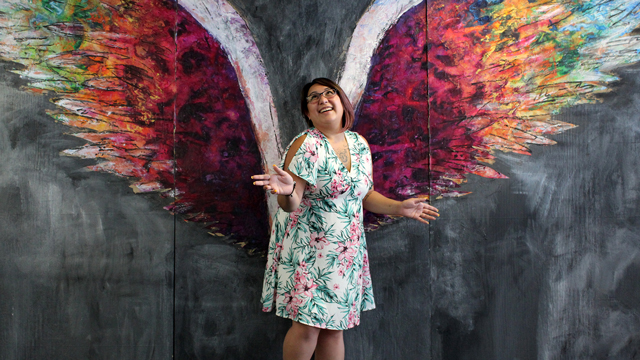 Vanessa McCarron poses in front of a colorful wall mural of angelic colorful wings.